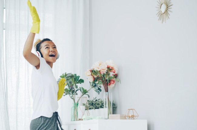 Pretty African American housewife is dusting the furniture and enjoying music wearing wireless headphones, dancing and singing. Happy young woman is having fun and doing housework.